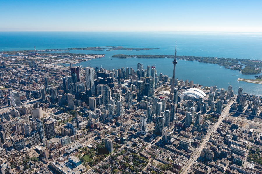 Aerial view of downtown Toronto on a clear day, showing a dense cluster of high-rise buildings, the CN Tower, the Rogers Centre, and Lake Ontario stretching out in the background.