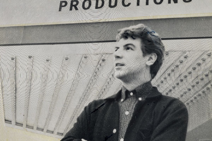 A duotone image in black and yellow showing a young man with his arms crossed, looking slightly upward with a sense of optimism. A sign on the wall behind him reads "Watch for upcoming productions".