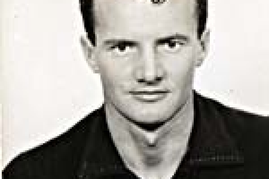 A black and white photo of Walter Grealis wearing a black button up shirt. He has short styled hair with a curly lock in the front. 