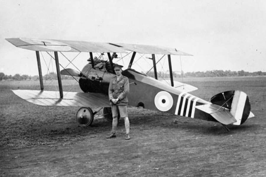 A vintage black‑and‑white photograph of a World War I–era biplane parked on an open airfield. The aircraft has roundel markings on its fuselage and bold striped patterns on the tail. A uniformed pilot stands in front of the plane, hands clasped, looking toward the camera. The background shows flat terrain with trees along the horizon.