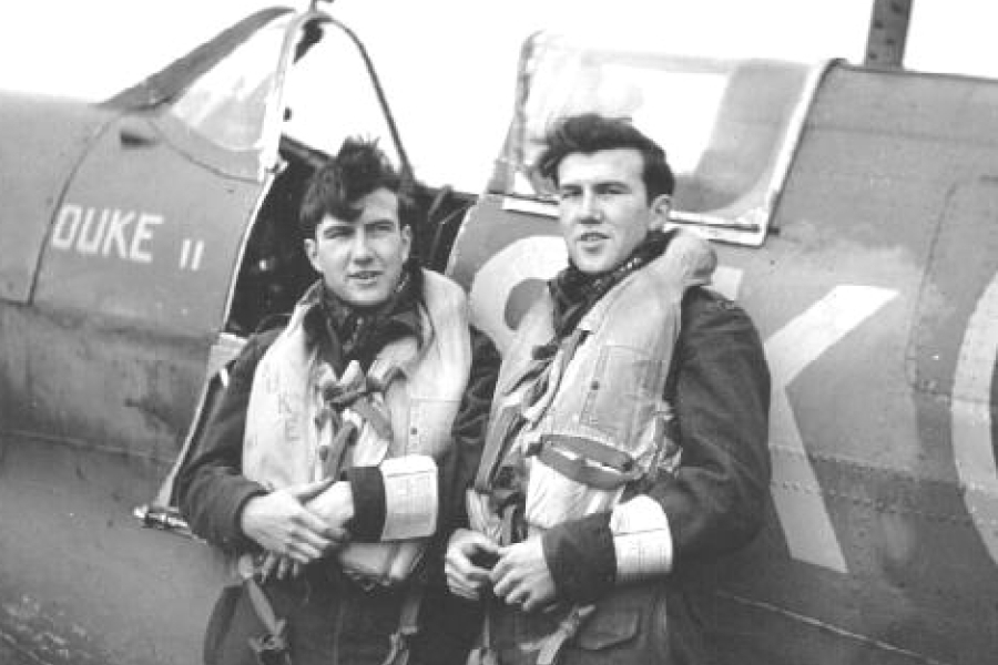 Black-and-white photograph of the identical twins Bruce and Duke Warren standing together beside a fighter aircraft marked “DUKE II.” Both wear flight jackets and life vests, with the cockpit and nose of the plane visible behind them, suggesting preparation for or return from a flight.