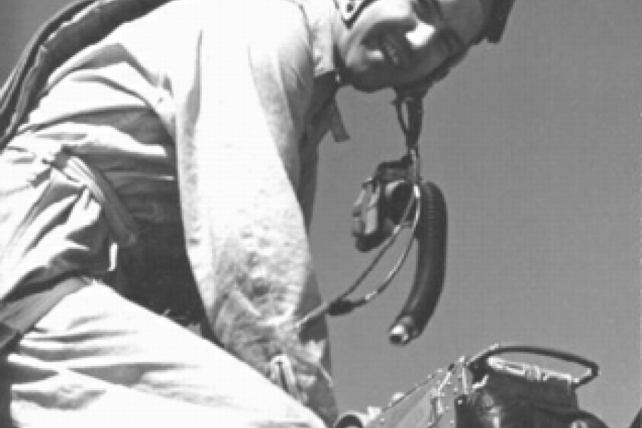 Black-and-white photograph of Bruce Warren wearing a flight suit, helmet, and goggles, crouched beside an aircraft while working with equipment on the fuselage. The angle is close and slightly below, emphasizing the technical details of the gear and the aircraft surface.