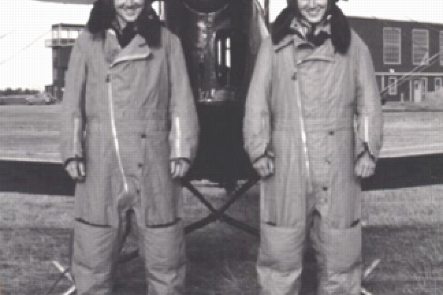 Black-and-white photograph of the twin aircrew members, Bruce and Duke Warren, standing side by side on a grassy airfield in front of a small military aircraft. They are wearing heavy flight suits and boots. They are smiling and posing in front of the plane with hangar buildings visible in the background.