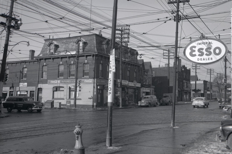 Black‑and‑white photograph of a city street intersection in 1957, showing brick commercial buildings, overhead streetcar wires, parked cars, and storefront signage. An “Imperial Esso Dealer” sign stands on the sidewalk, with utility poles, a fire hydrant, and early automobiles visible along Parliament Street in Toronto.