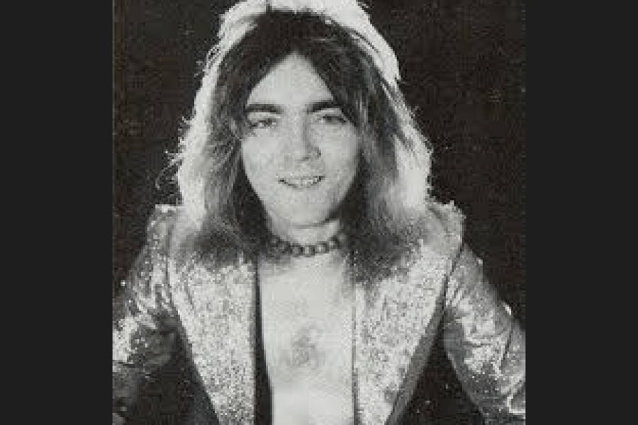 Black‑and‑white studio portrait of John Rutsey with long hair, wearing a patterned jacket over a light‑coloured shirt, posed against a dark background.