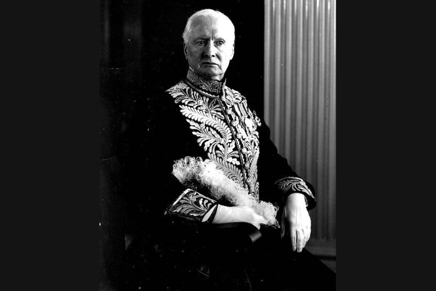 Black‑and‑white formal studio portrait of a person seated and wearing an ornate, embroidered ceremonial jacket with decorative braiding and medals. The individual rests one arm on a chair, with a feathered accessory across their lap, posed against a dark backdrop with a textured column visible to one side.