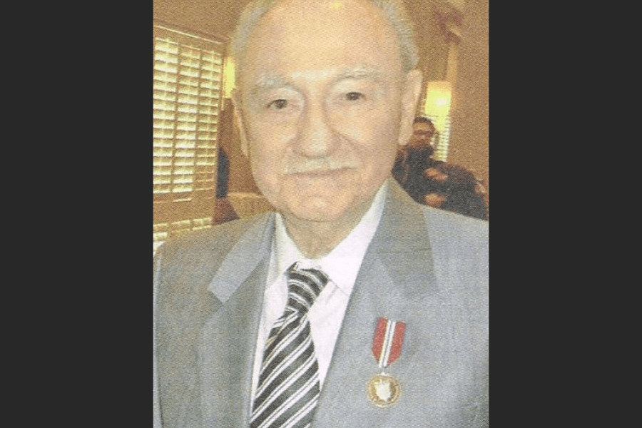 Formal portrait of a person wearing a light‑coloured suit and striped tie, with a military or service medal pinned to the left lapel. The photograph appears to be taken indoors, with window blinds and seated figures visible in the softly lit background.