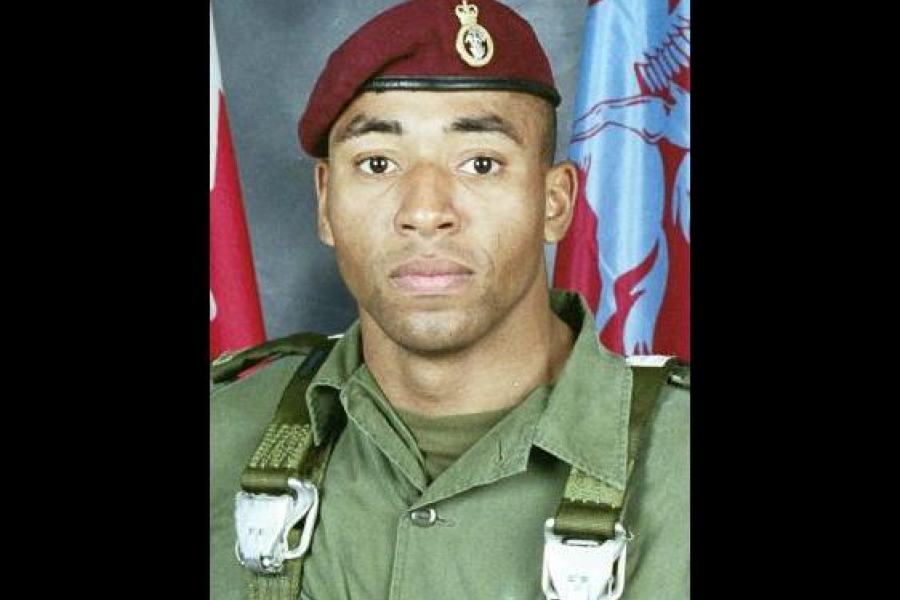 Portrait of Corporal Ainsworth Dyer in military uniform, wearing a maroon beret and standing in front of flags, photographed from the chest up.