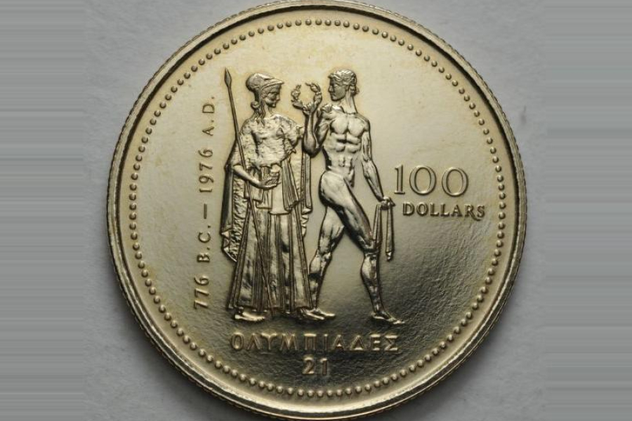 A close-up photograph of a gold commemorative coin featuring two classical figures standing in the center—one draped and holding a staff, the other nude and athletic, holding a wreath. The inscriptions include “776 B.C. – 1976 A.D.” along the left side, “100 DOLLARS” on the right, and Greek lettering along the bottom. The coin rests on a light, textured background.