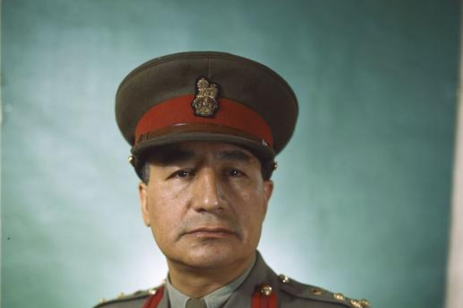 Studio portrait of a military officer wearing a formal uniform with a peaked cap, medal ribbons, and aviation wings, posed against a plain teal backdrop and shown from the waist up.