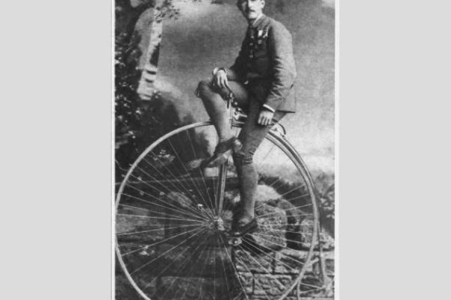 Historic black‑and‑white photograph of a man wearing a suit and tie, posed seated on a large high‑wheel bicycle in an outdoor studio setting with a painted backdrop of trees and foliage.