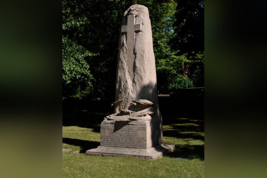 Empress of Ireland Memorial, a tall, rough‑hewn stone monument carved with a prominent cross near the top, standing on a square inscribed base on a grassy lawn, with mature trees forming a shaded background.