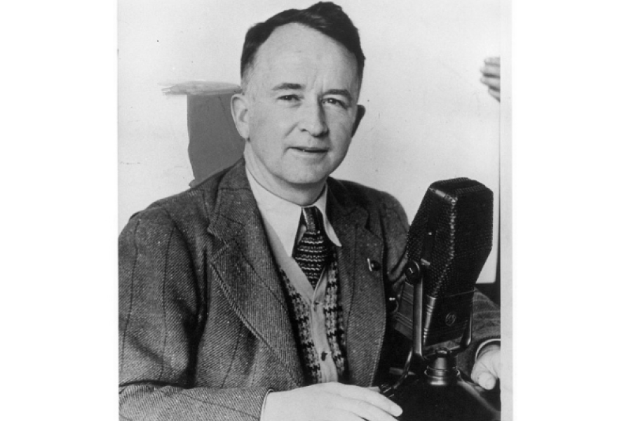 Black‑and‑white archival photograph of a person seated at a desk, wearing a suit jacket, patterned tie, and vest, speaking into a large tabletop broadcast microphone, with one hand resting on the desk in what appears to be a radio studio setting.
