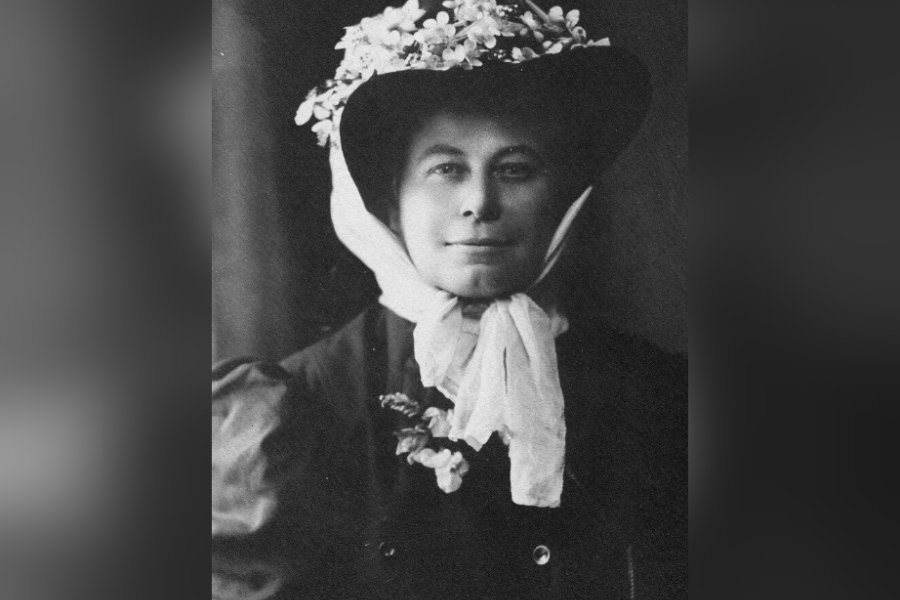 Black‑and‑white portrait of Alice Eastwood wearing a dark coat and a wide‑brimmed hat adorned with flowers, with a light scarf tied at the neck, photographed against a dark background.