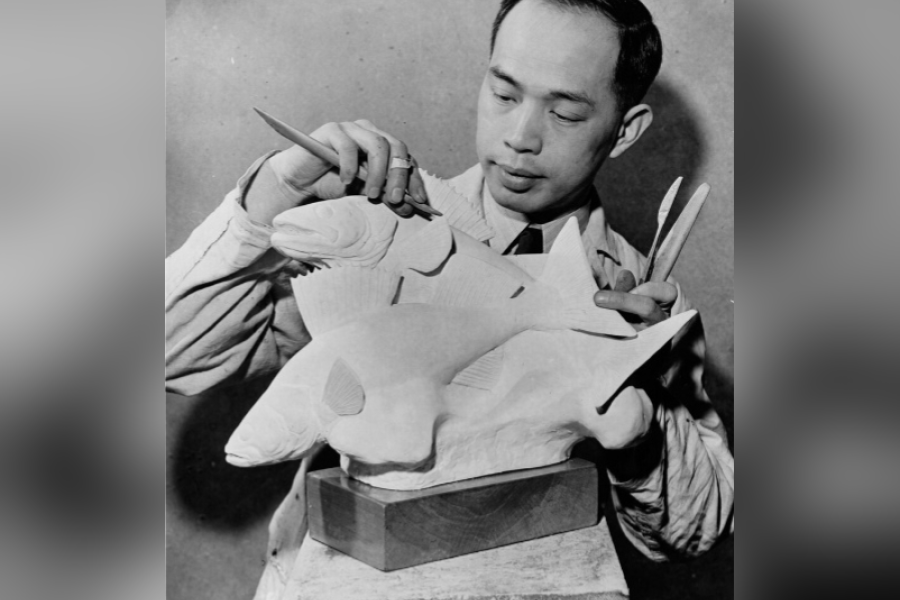 Black‑and‑white photograph of Sing Hoo Yuen working on a large fish sculpture mounted on a pedestal, holding carving tools in both hands while shaping the fins and body, wearing a work coat in a studio setting.