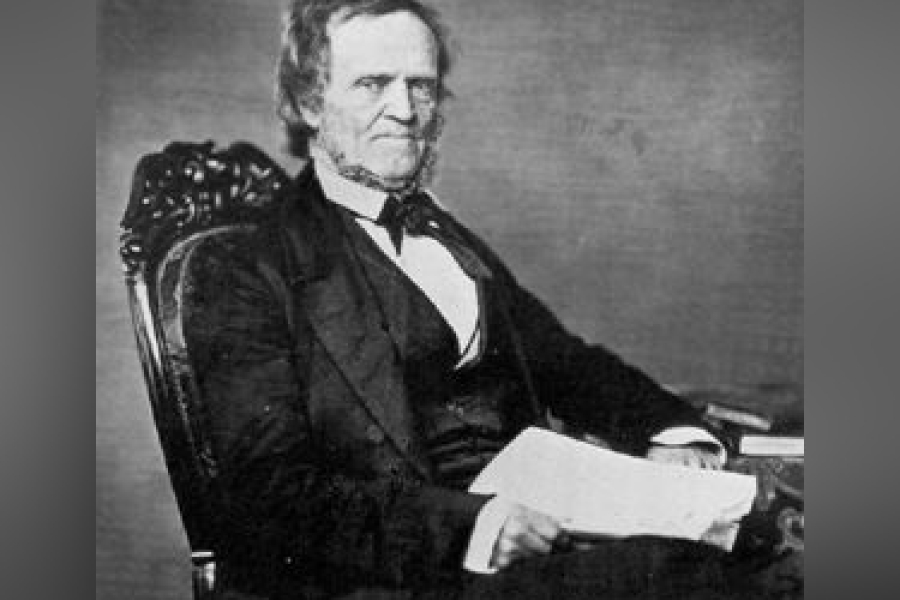 Black‑and‑white seated portrait of William Lyon Mackenzie wearing a dark suit, waistcoat, and bow tie, holding papers while resting one arm on a table, seated in an ornate chair against a plain studio backdrop.
