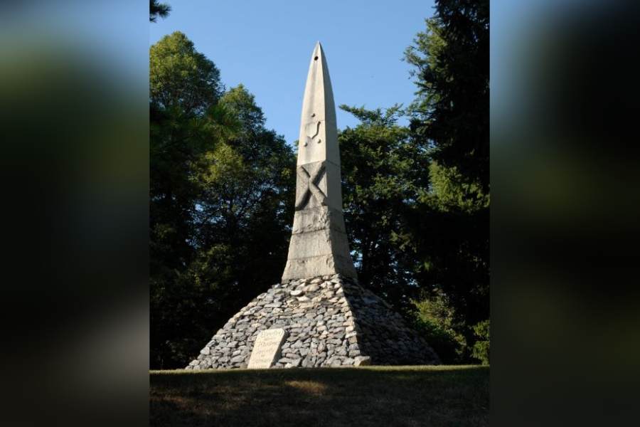 St. Andrew’s Society Cairn, a tall stone obelisk rising from a circular base of stacked grey stones with an inscribed plaque, set on a grassy slope and framed by mature trees beneath a clear blue sky.