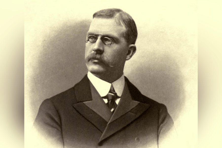 Portrait of J. B. Tyrrell wearing a dark, structured suit jacket with wide lapels, a high‑collared shirt, and a patterned tie, shown from the chest up against a softly shaded oval studio background.