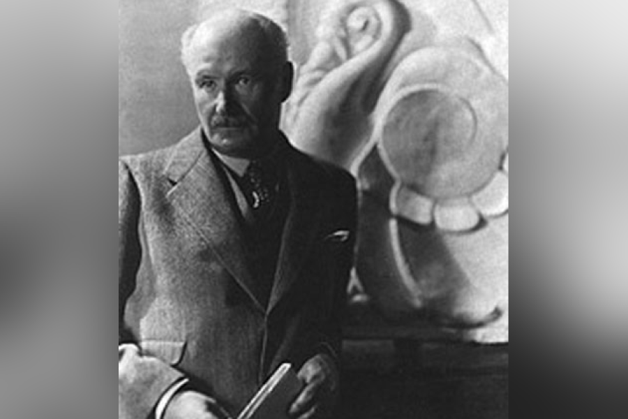 Black‑and‑white photograph of John M. Lyle standing in front of a large sculptural relief, wearing a suit and tie and holding carving tools in his hands, suggesting an artist or architectural studio setting.