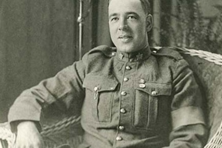Black‑and‑white photograph of Sidney Lambert seated in a wicker chair, wearing a First World War military uniform with a buttoned tunic and insignia, resting one arm on the chair, with an indoor setting and standing lamp visible behind him.