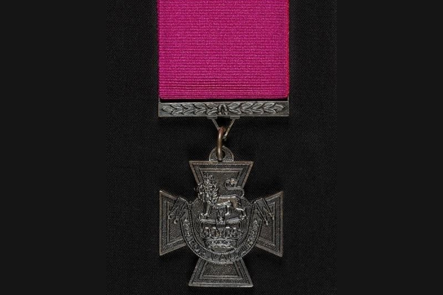 A detailed photograph of a Victoria Cross medal suspended from a deep magenta ribbon. The bronze cross features a lion standing atop a royal crown at its center, with an ornate scroll below. The medal hangs against a dark, textured background