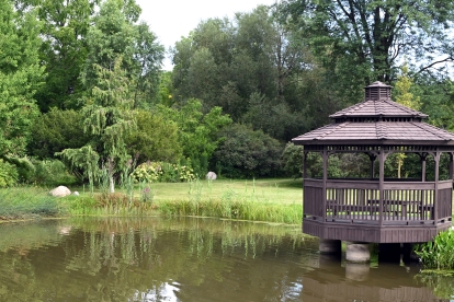 A wooden gazebo on stilts extends over the edge of a calm pond, surrounded by lush green grass and dense trees. The scene includes a variety of foliage, such as a large weeping willow on the left and tall leafy trees in the background, creating a peaceful park-like setting.