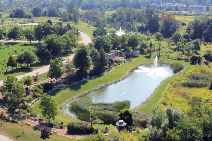 Aerial view of a landscaped cemetery featuring green lawns with rows of headstones, winding paved paths, and a large pond with a fountain at its center. The area is surrounded by trees and shrubs, with open fields and distant residential buildings visible in the background.