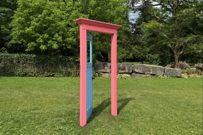 Pink decorative doorframe standing on a grassy lawn, with trees, stone benches, and greenery in the background.
