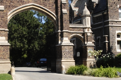 Historic stone gates of Mount Pleasant Cemetery in Toronto, featuring a large central arch flanked by smaller arches and towers, surrounded by manicured gardens with colorful flowers and greenery.