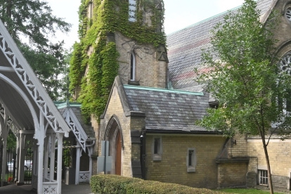 Entrance to Necropolis Cemetery featuring a white Gothic-style archway with intricate wooden detailing, leading to a historic stone chapel partially covered in ivy. The chapel has tall arched windows and a slate roof, surrounded by lush green trees and a stone monument in the foreground.