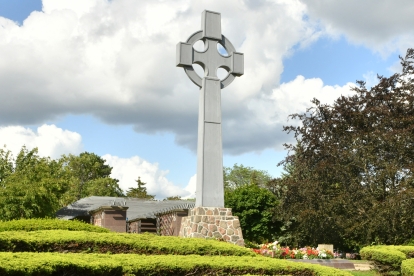 Large Celtic cross monument standing on a stone base, surrounded by manicured green hedges and trees under a partly cloudy sky.