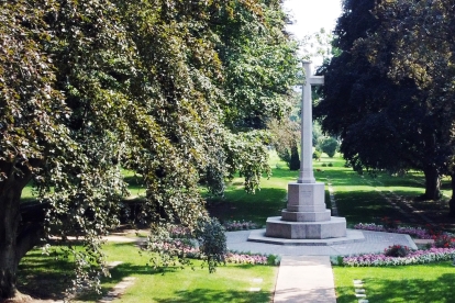 Stone monument with a tall cross at the center of a landscaped garden, surrounded by green grass, flower beds, and large trees providing shade. Pathways lead to the monument from multiple directions under bright daylight.