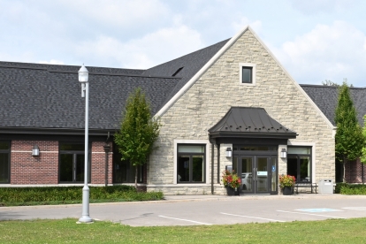 Single-story building with a stone and brick exterior, dark shingled roof, and multiple peaked gables. The building has large windows and a central entrance with potted plants. Several white vehicles are parked in front on a paved lot, surrounded by green grass, trees, and lampposts under a partly cloudy sky.