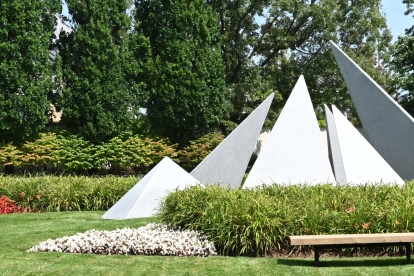 Outdoor memorial with several large white triangular stone structures arranged in a landscaped garden, surrounded by green grass, colorful flower beds in red and white, and benches. Tall trees and shrubs form the background.