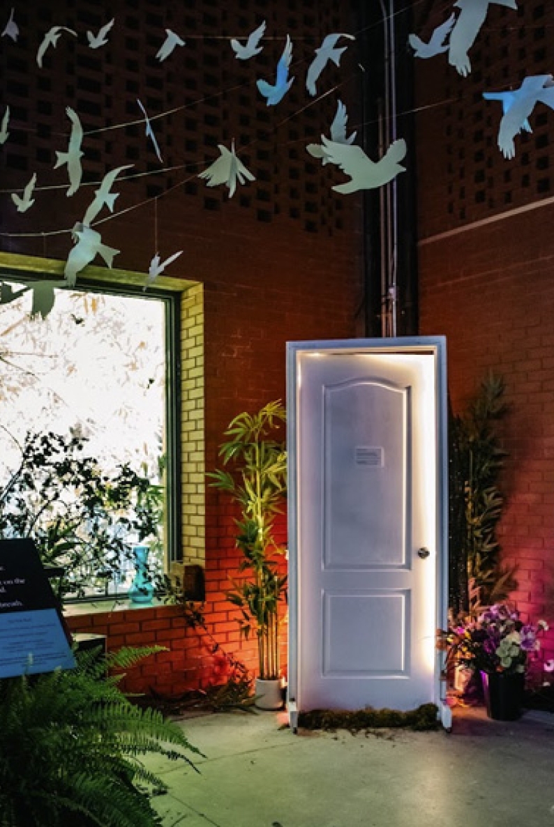 A lit white door stands in the center of an indoor art installation, surrounded by plants and colorful accent lighting. Paper birds hang from the ceiling on thin wires, appearing to fly overhead. A large window on the left and a small sign near the plants complete the scene.