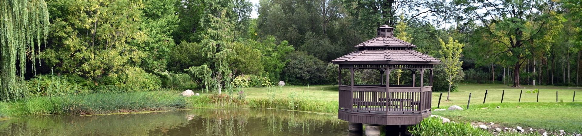 A wooden gazebo on stilts extends over the edge of a calm pond, surrounded by lush green grass and dense trees. The scene includes a variety of foliage, such as a large weeping willow on the left and tall leafy trees in the background, creating a peaceful park-like setting.