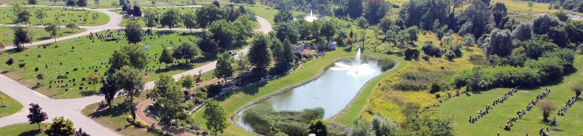 Aerial view of a landscaped cemetery featuring green lawns with rows of headstones, winding paved paths, and a large pond with a fountain at its center. The area is surrounded by trees and shrubs, with open fields and distant residential buildings visible in the background.
