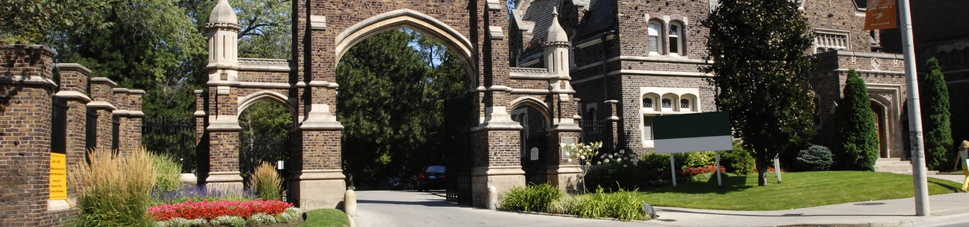 Historic stone gates of Mount Pleasant Cemetery in Toronto, featuring a large central arch flanked by smaller arches and towers, surrounded by manicured gardens with colorful flowers and greenery.