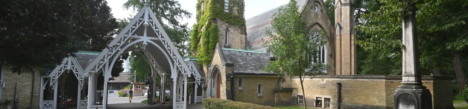 Entrance to Necropolis Cemetery featuring a white Gothic-style archway with intricate wooden detailing, leading to a historic stone chapel partially covered in ivy. The chapel has tall arched windows and a slate roof, surrounded by lush green trees and a stone monument in the foreground.