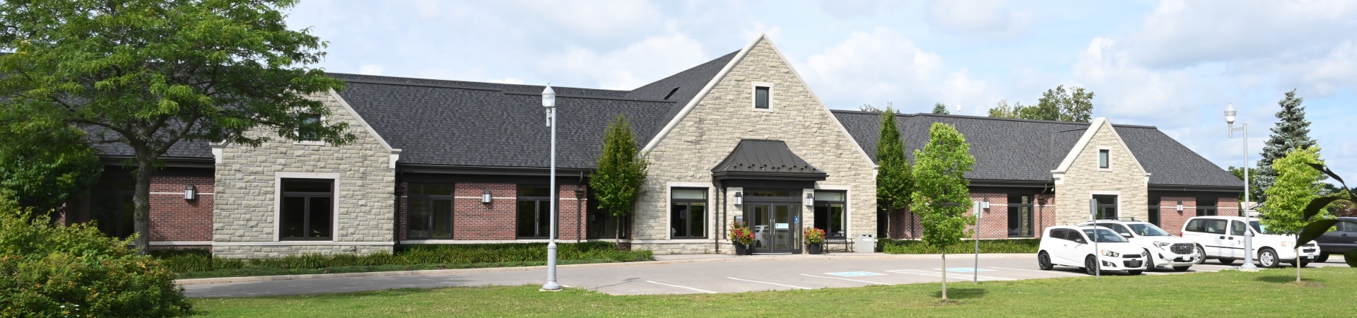 Single-story building with a stone and brick exterior, dark shingled roof, and multiple peaked gables. The building has large windows and a central entrance with potted plants. Several white vehicles are parked in front on a paved lot, surrounded by green grass, trees, and lampposts under a partly cloudy sky.
