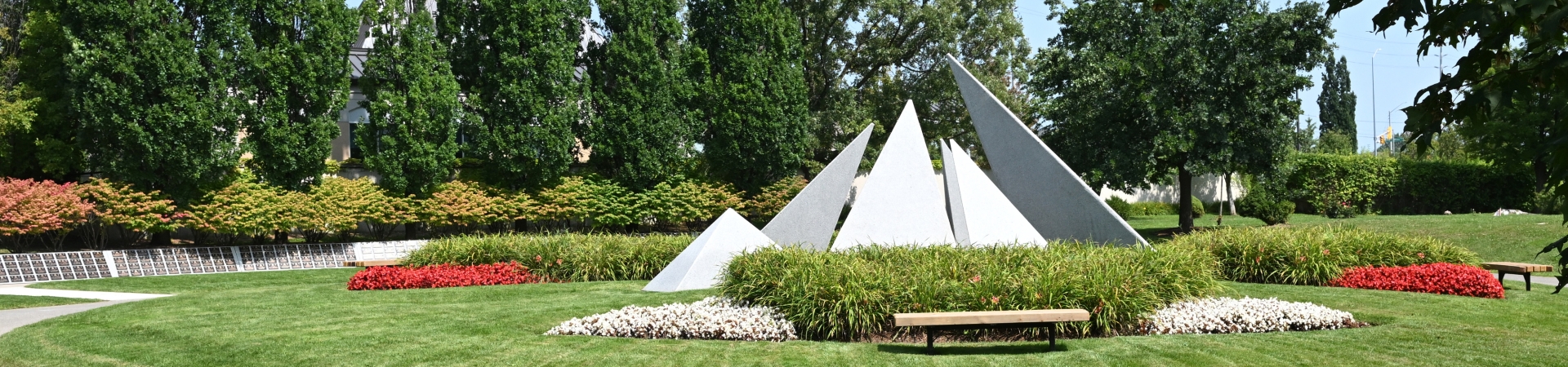 Outdoor memorial with several large white triangular stone structures arranged in a landscaped garden, surrounded by green grass, colorful flower beds in red and white, and benches. Tall trees and shrubs form the background.